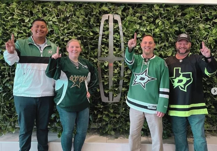 Four people stand in front of a wall covered in plants. The lincoln car brand emblem is in the center of the wall. All four people are wearing green sports jerseys for the Dallas Stars sports team and smiling.